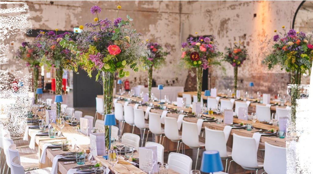 Lange Banketttische mit weißen Stühlen und hohen floralen Tafelaufsätzen füllen einen rustikal dekorierten Veranstaltungssaal.