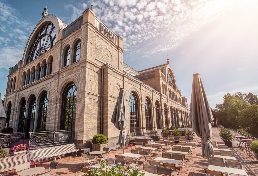 Historisches Backsteingebäude mit großen Bogenfenstern und einer Außenterrasse mit Tischen, geschlossenen Sonnenschirmen und dem einladenden Ambiente des Catering im Dank Augusta.