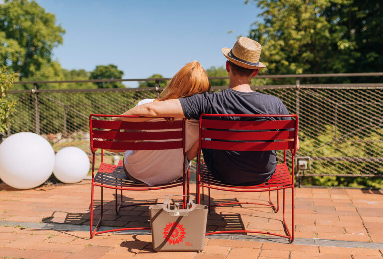 Eng zusammensitzt ein Pärchen auf roten Stühlen im Freien und genießt einen sonnigen Tag mit leckeren Leckereien von Catering im Dank Augusta.