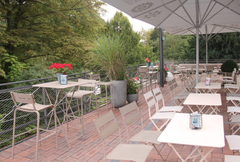Außenterrasse mit leeren Tischen und Stühlen, Topfpflanzen und großen Sonnenschirmen, umgeben von Grün – eine einladende Umgebung, perfekt für Catering im Dank Augusta.