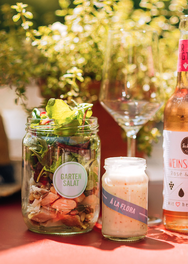 Ein Glas Gartensalat, ein kleines Glas Dressing, ein Weinglas und eine Flasche auf einem Tisch mit Pflanzen dahinter – perfekt gestylt für Catering im Dank Augusta.