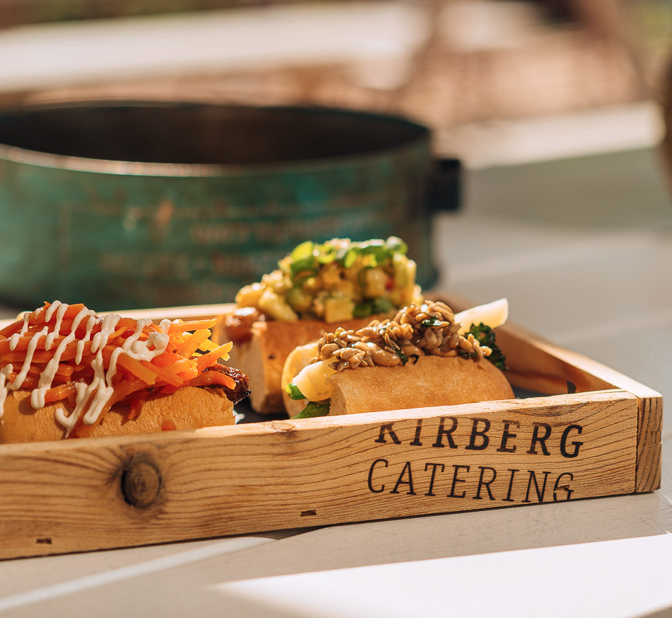 Drei Gourmet-Hotdogs mit verschiedenen bunten Toppings auf einem Holztablett mit der Aufschrift „Kirberg Catering“, perfekt für Catering im Dank Augusta.