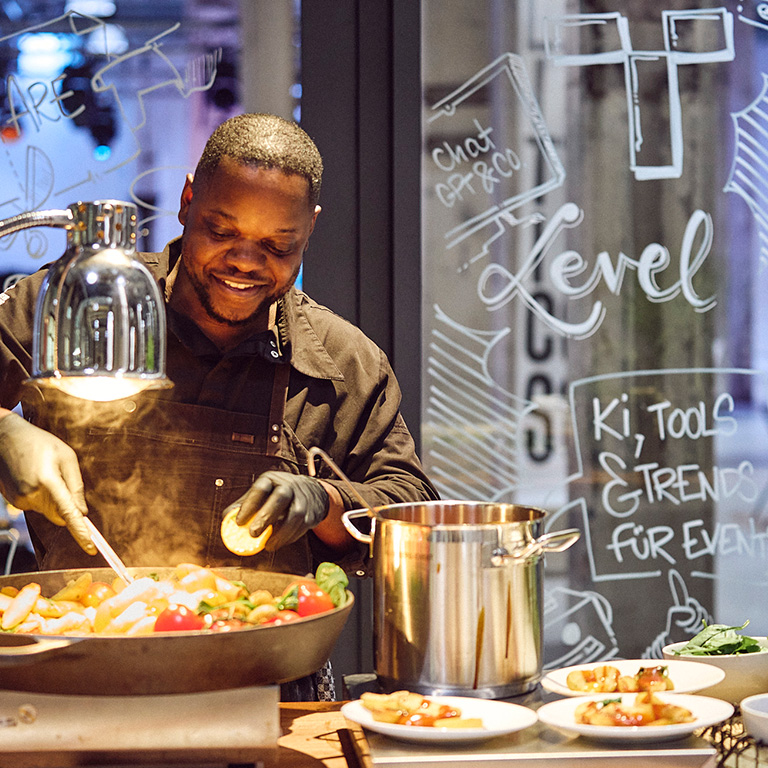 Ein Kirberg-Koch rührt ein Kartoffelgericht, das geschmackvoll mit Tomaten und grünem Gemüse in einer Pfanne zubereitet wird.
