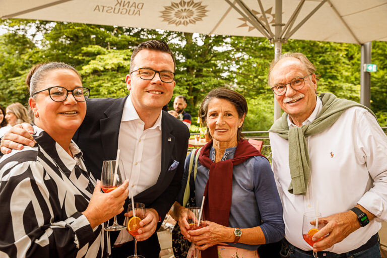 Vier lächelnde Erwachsene mit Getränken posieren gemeinsam im Freien unter einem Sonnenschirm bei einer gesellschaftlichen Veranstaltung.