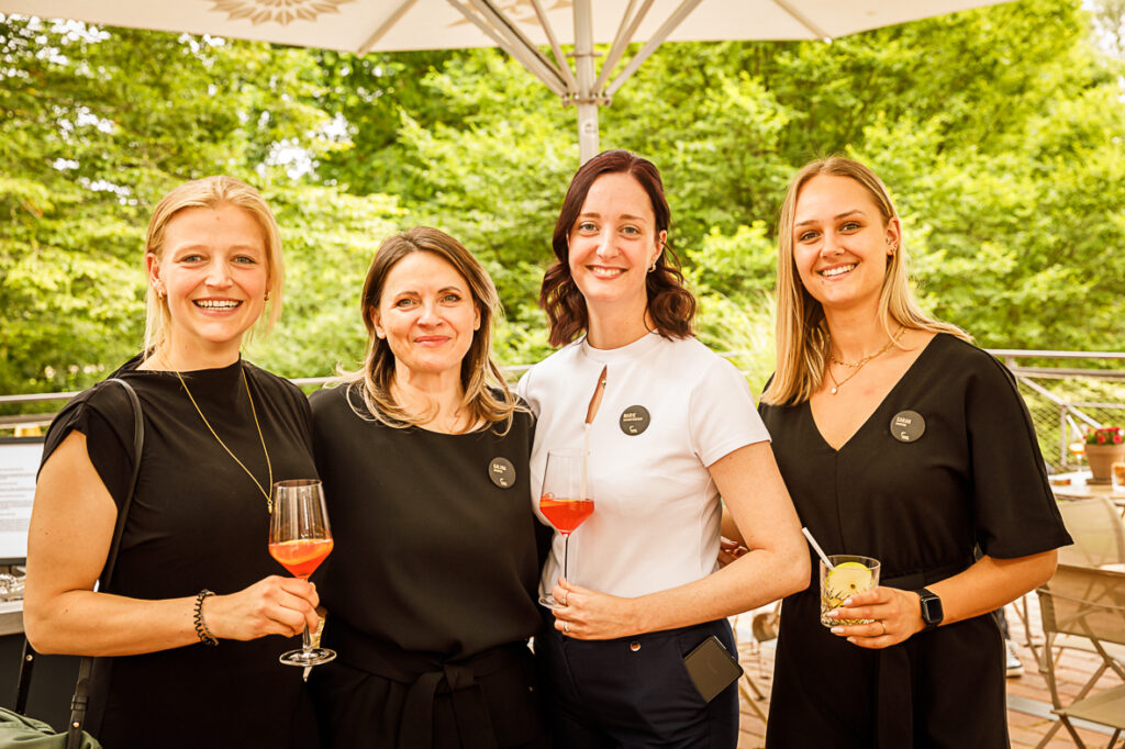 Vier lächelnde Frauen im Freien, jede mit einem Getränk in der Hand, stehen zusammen unter einem großen Sonnenschirm.