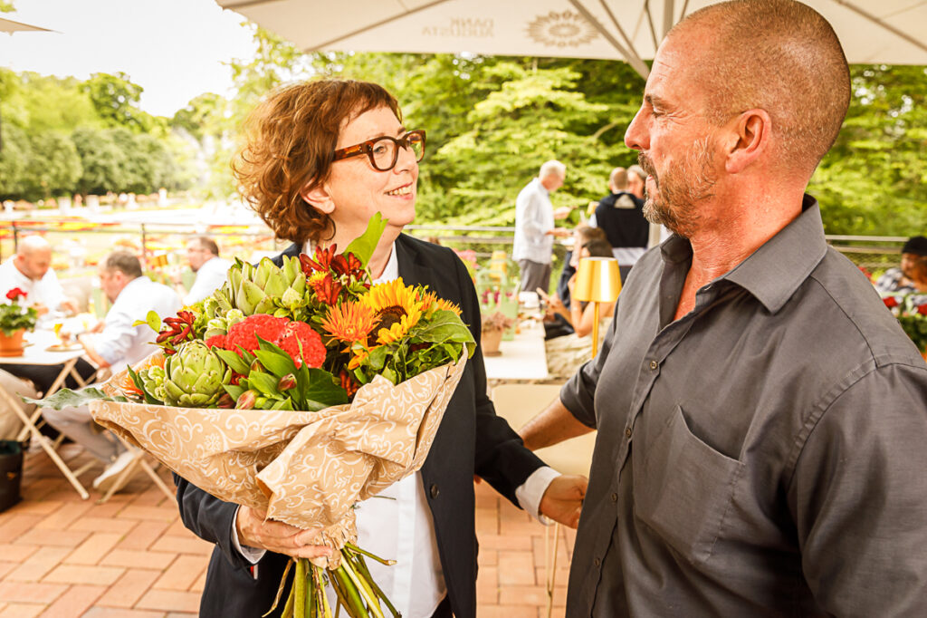 Eine Frau mit einem Blumenstrauß in der Hand lächelt einen Mann bei einem geselligen Beisammensein im Freien an.