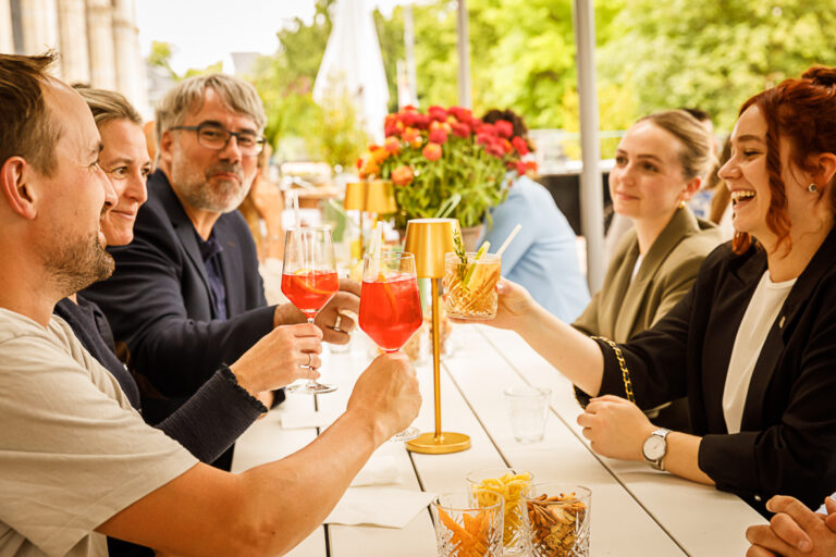 Fünf Personen stoßen an und lächeln, während sie mit Getränken und Snacks an einem Tisch im Freien sitzen.