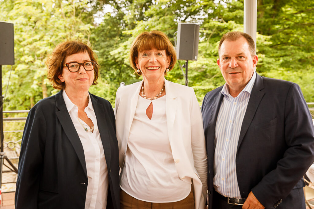 Drei Erwachsene in Geschäftskleidung stehen lächelnd im Freien, im Hintergrund Grün.