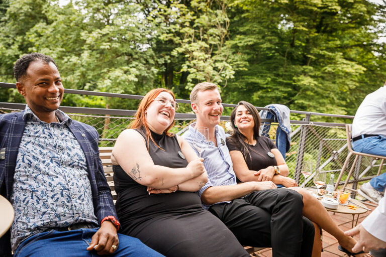 Vier Menschen sitzen lächelnd und lachend draußen auf einer Terrasse, im Hintergrund sind Bäume zu sehen.
