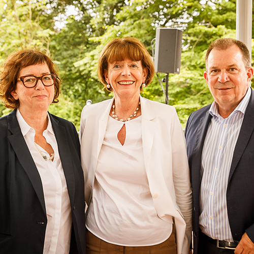 Drei Erwachsene in Geschäftskleidung stehen lächelnd im Freien, im Hintergrund Grün.