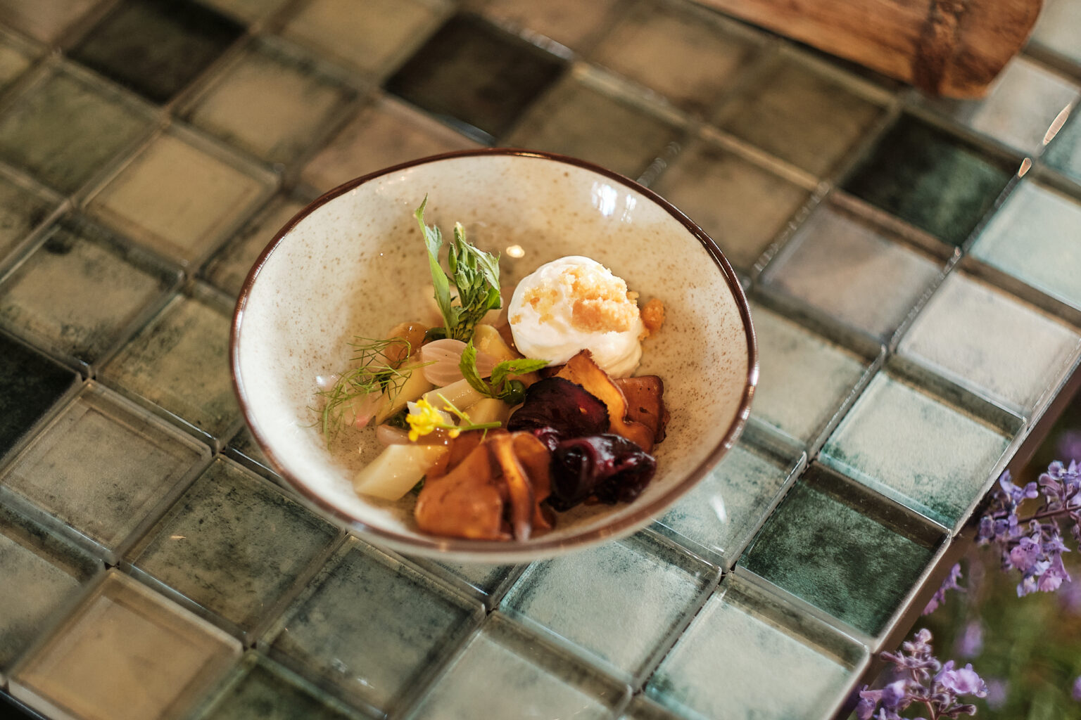 Eine Schale mit gebratenem Gemüse, Kräutern und einem Klecks Sahne auf einem gekachelten Glastisch.