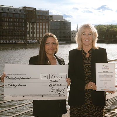 Zwei Frauen stehen an einem Fluss, halten einen großen Scheck und eine Urkunde für Nachhaltigkeit im Catering in der Hand und lächeln in die Kamera.