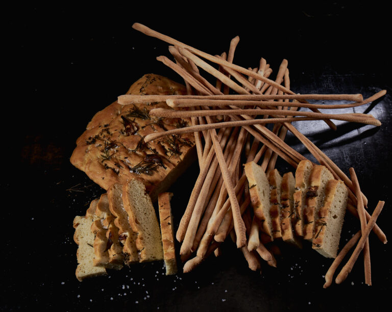 In Scheiben geschnittene Focaccia und lange Brotstangen, die auf einer dunklen Fläche mit ein paar Salzkörnern angerichtet sind.