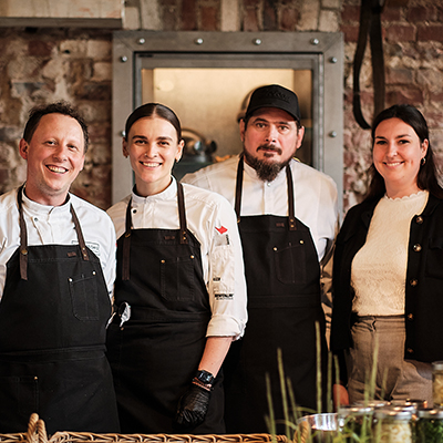 Vier Personen, darunter drei Köche in schwarzen Schürzen, stehen lächelnd zusammen in einer rustikalen Küche, die Nachhaltigkeit im Catering unterstreicht.