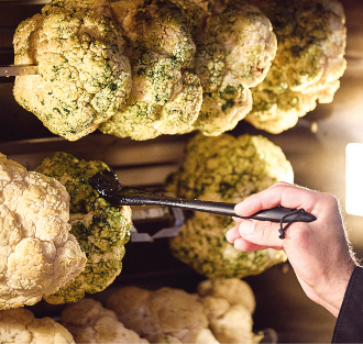 Eine Hand pinselt Gewürze auf ganze Blumenkohlköpfe, die in einer Rotisserie gebraten werden. Dies unterstreicht die Verwendung regionaler Produkte im Eventcatering für ein frisches und geschmackvolles Erlebnis.