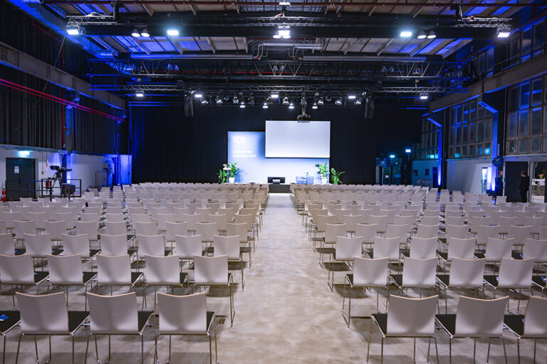 Ein leerer Konferenzsaal mit weißen Stuhlreihen vor einer Bühne mit Leinwand und Pflanzen.