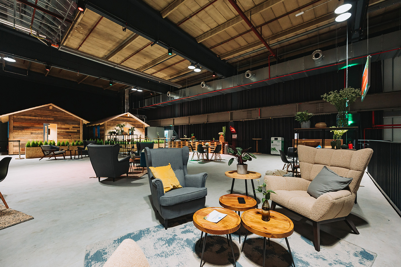 Moderne Indoor-Lounge mit gemütlichen Sitzgelegenheiten, Holztischen und hüttenähnlichen Strukturen im Hintergrund.