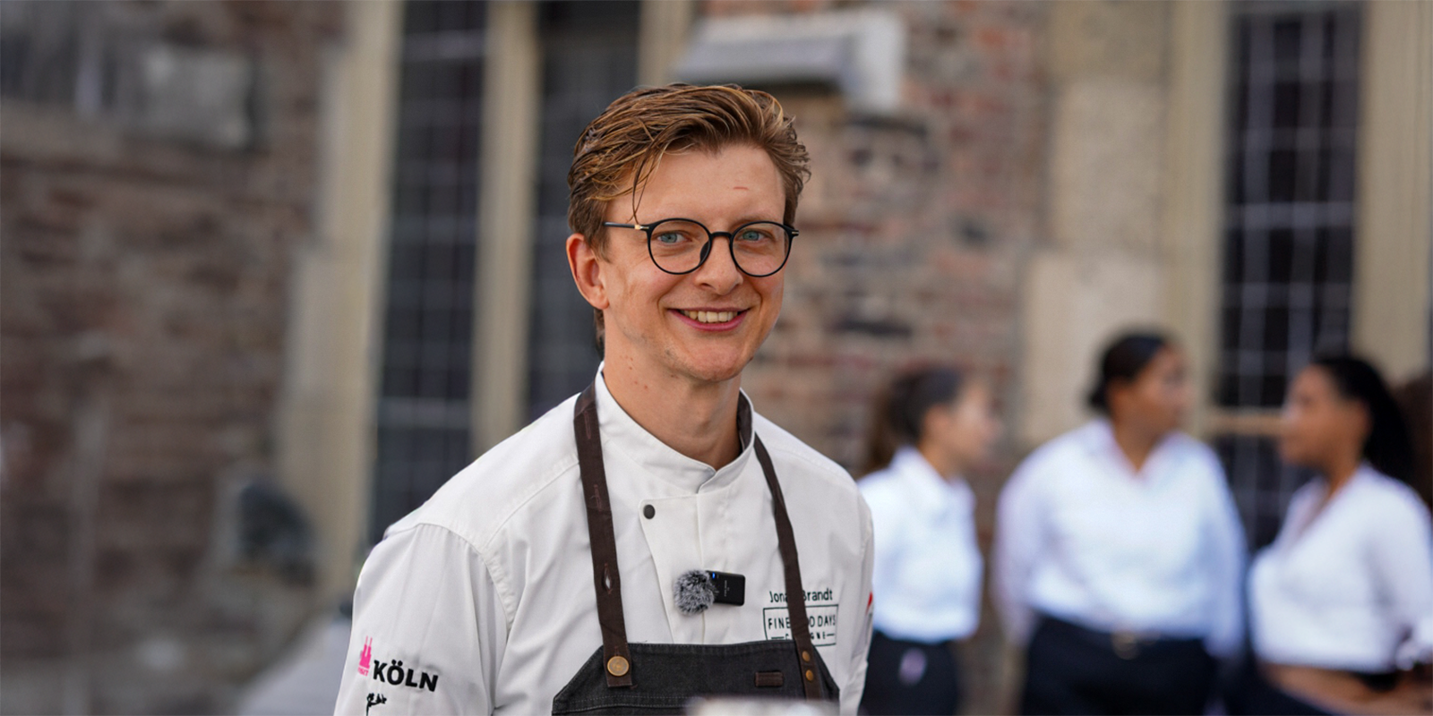 Ein lächelnder Koch mit Brille und Uniform steht im Freien, vier Personen in weißen Hemden sind unscharf im Hintergrund zu sehen.