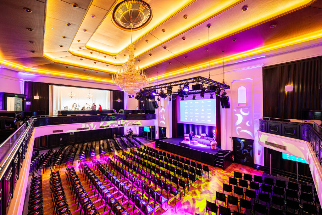 Ballsaal in der Eventlocation Curio Haus in Köln mit Stuhlreihen vor einer Bühne, lila Beleuchtung und einem großen Kronleuchter über dem Kopf.