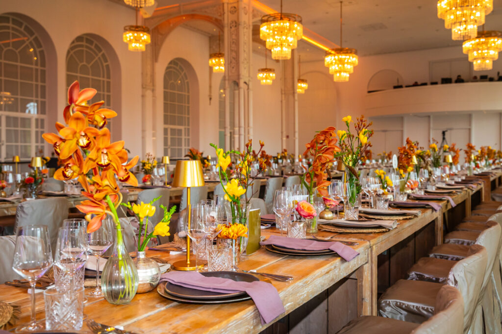 Eleganter Bankettsaal mit langen Holztischen, gedeckt mit Blumen, Glaswaren und goldenen Akzenten unter Kronleuchtern.