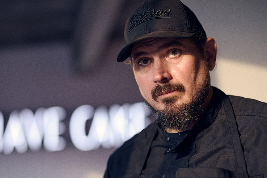 Ein bärtiger Mann mit schwarzer Mütze und Jacke blickt in einer schwach beleuchteten Umgebung in die Kamera und fängt eine urbane Stimmung ein, die von Philara 14 Places inspiriert ist.