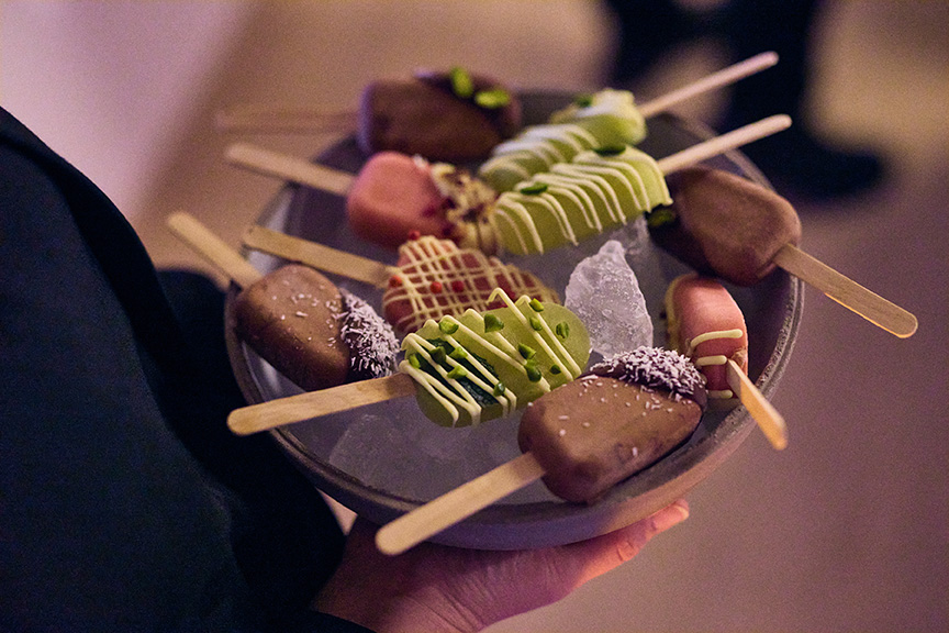 Eine Schale mit verschiedenen bunten Eisstielen, die von einer dunkel gekleideten Person auf Eis gehalten wird, fängt den spielerischen Geist von Philara 14 Places ein.