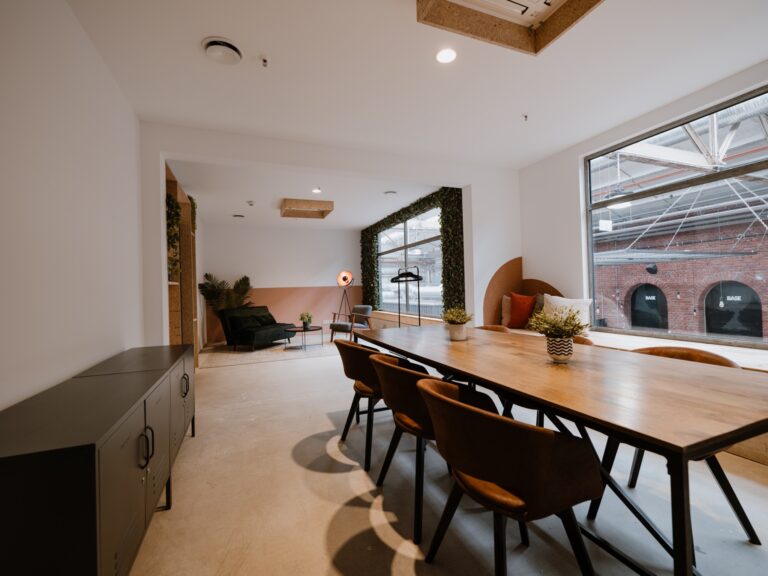 Moderner Büroraum mit langem Holztisch, Stühlen, Pflanzen, Lounge-Bereich und großen Fenstern.