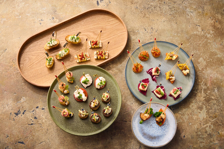 Verschiedene mundgerechte Häppchen auf vier Tellern, ordentlich auf einer strukturierten braunen Oberfläche angeordnet - perfekt zum Probieren von Geschmacksrichtungen, die vom lebendigen Geist von Philara 14 Places inspiriert sind.