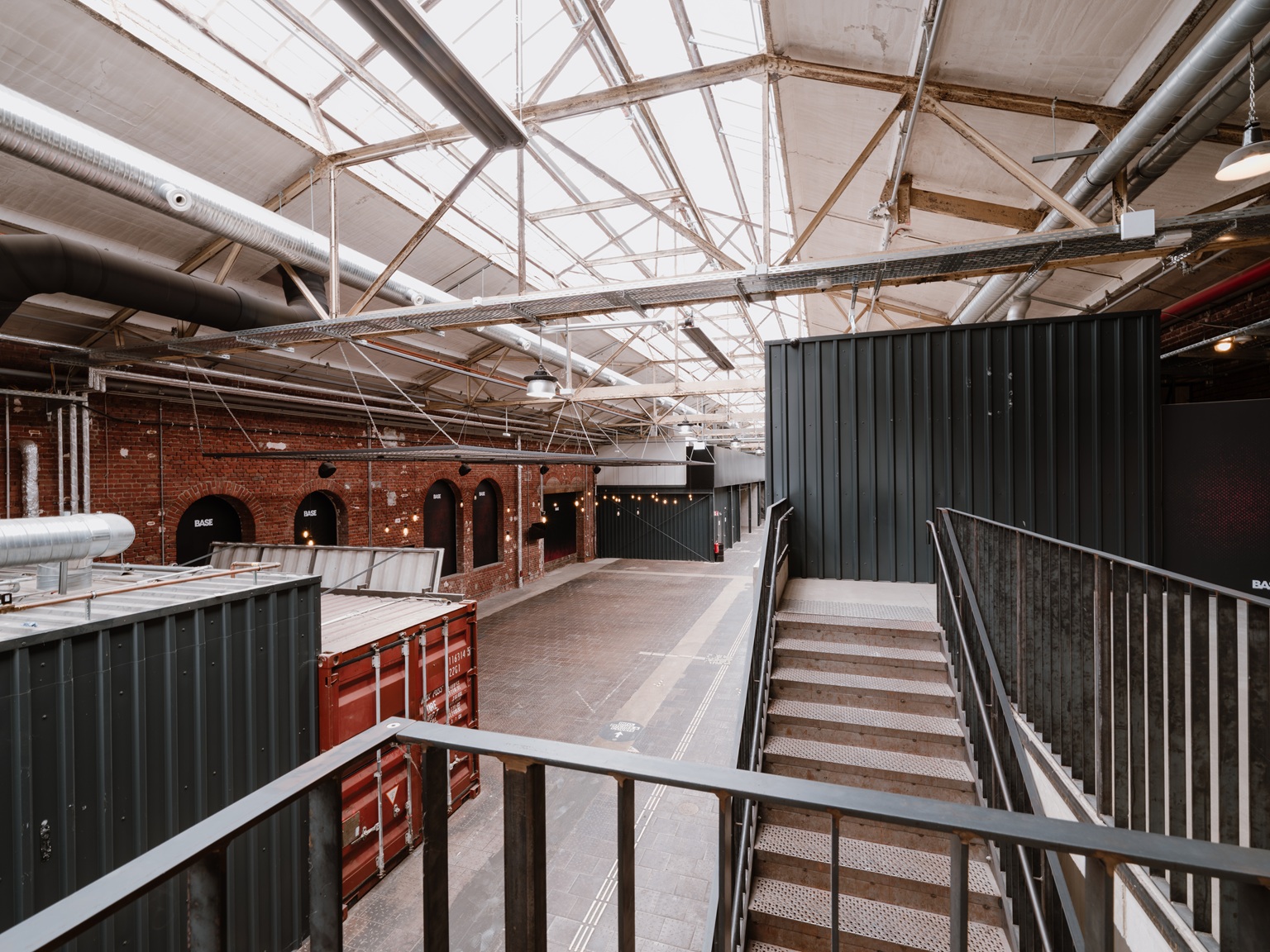 Industrielles Interieur mit Metalltreppen, Schiffscontainern, Ziegelwänden und sichtbaren Balken unter einem Glasdach.