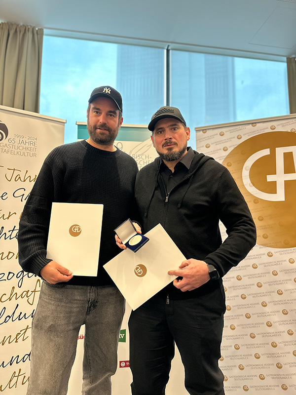 Zwei Männer mit Urkunden und einer Auszeichnung für 14 Plätze von Philara, die bei einer Veranstaltung in einer Halle vor Bannern stehen.