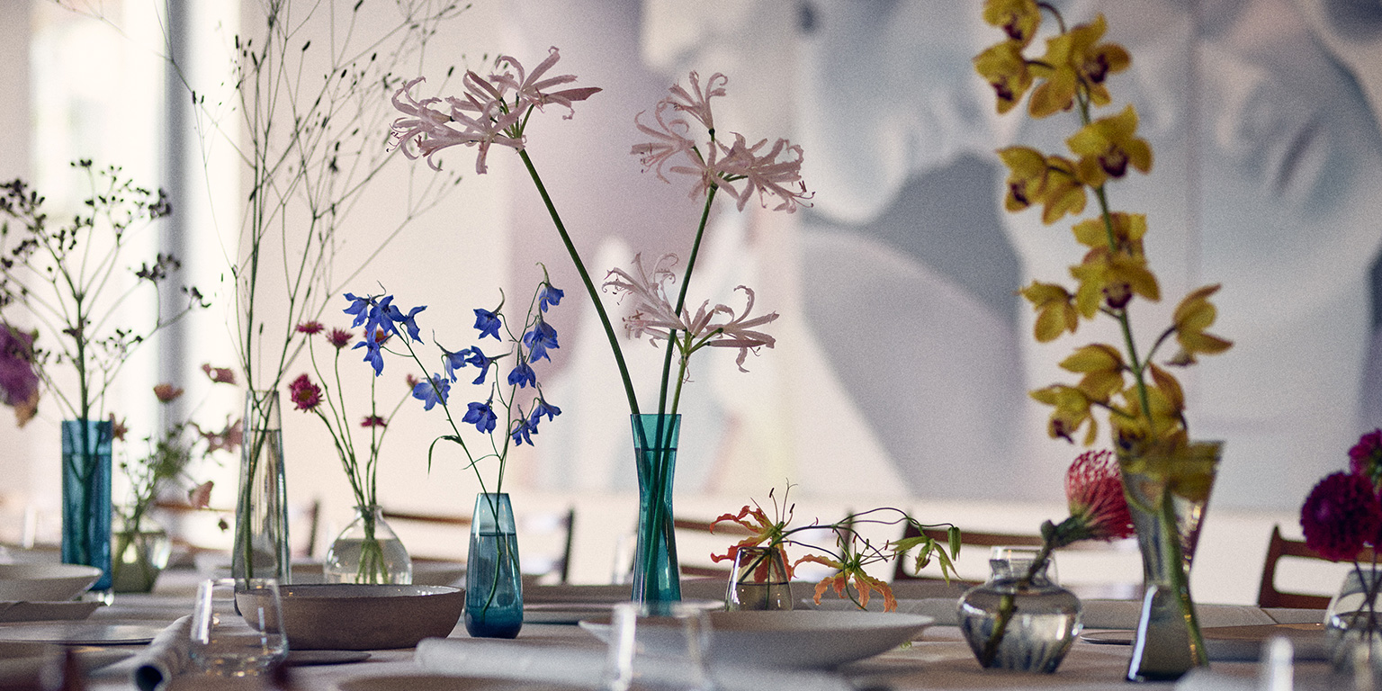 Verschiedene bunte Blumen in Glasvasen, die auf einem Tisch in einer sanft beleuchteten, eleganten Umgebung arrangiert sind, vermitteln den heiteren Charme von Philara 14 Places.