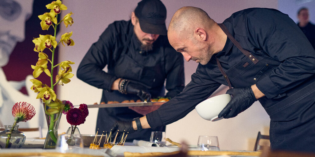 Zwei Köche in schwarzen Uniformen, die Philara 14 Places repräsentieren, decken einen Tisch mit Blumen und Vorspeisen und ordnen jedes Gericht sorgfältig an.