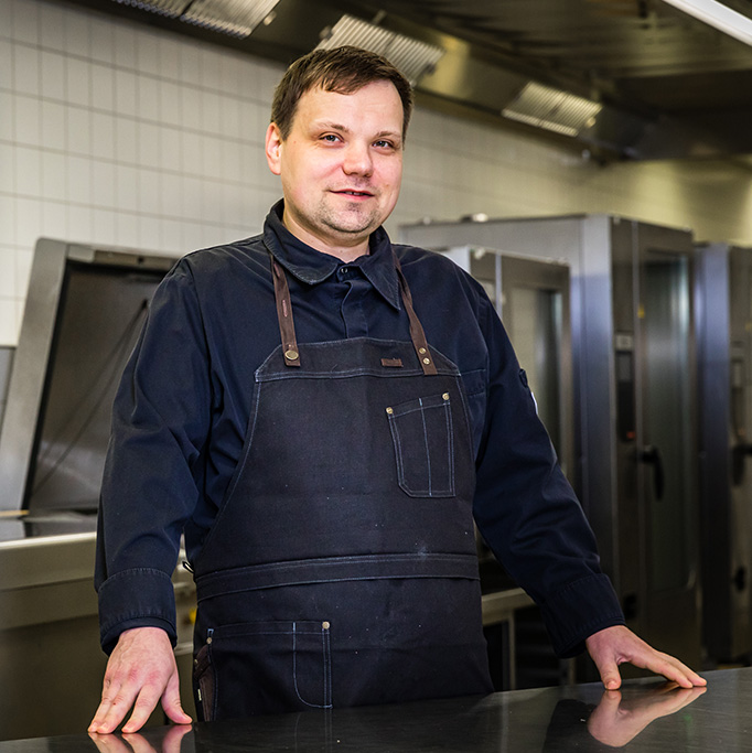 Ein Koch in einer dunklen Schürze steht lächelnd in einer professionellen Küche mit Edelstahlgeräten.