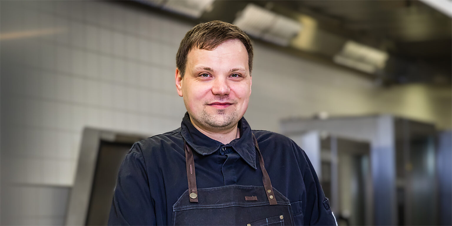 Ein Mann in einer dunklen Kochuniform steht lächelnd in einer Großküche.