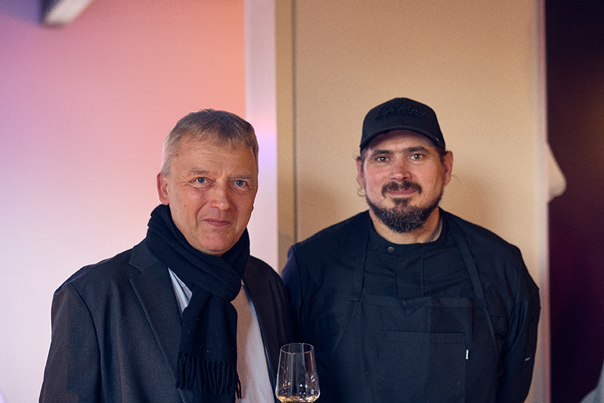 Zwei Männer stehen im Innenbereich von Philara 14 Places, einer hält ein Weinglas, beide schauen in die Kamera und lächeln sanft.