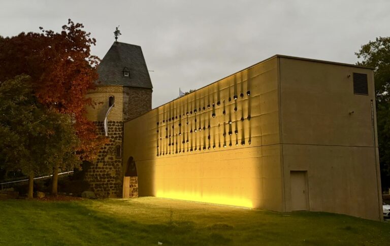 Ein modernes Gebäude mit gelben Lichtern steht neben dem historischen Sachsenturm Köln, dem Sitz von Kirberg Catering, umgeben von Bäumen in der Abenddämmerung.