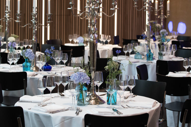 Elegant gedeckte Tische für eine Veranstaltung bei Catering in der BASE Köln - Eventlocation für Business, Kultur & private Feiern, mit blauen Blumen, Gläsern und silbernen Kandelabern auf weißen Tischtüchern.