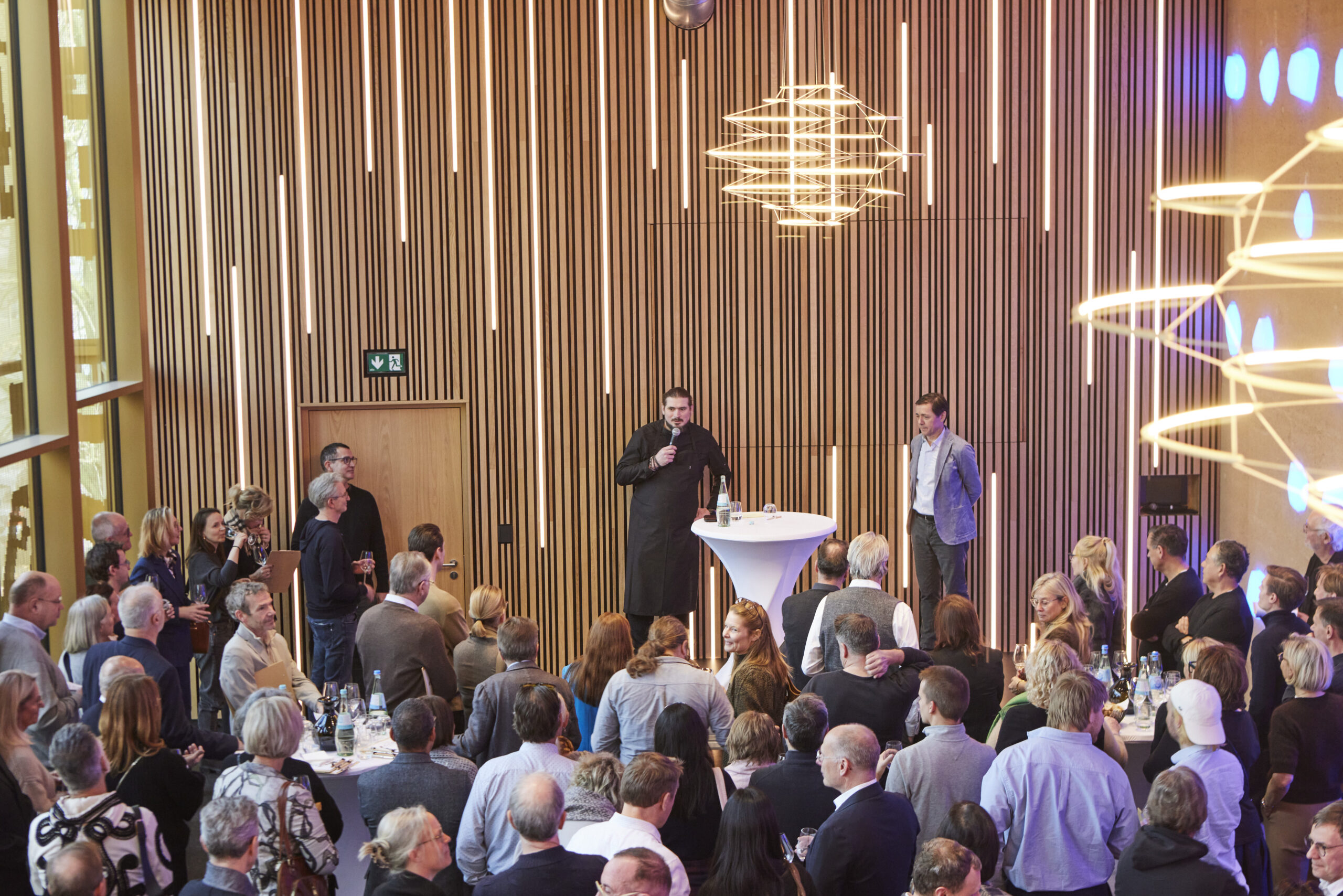 Eine große Gruppe hört einem Mann zu, der während einer Indoor-Veranstaltung im modernen, holzgetäfelten Raum des Kirberg Catering Sachsenturm Köln auf einem Podium spricht.