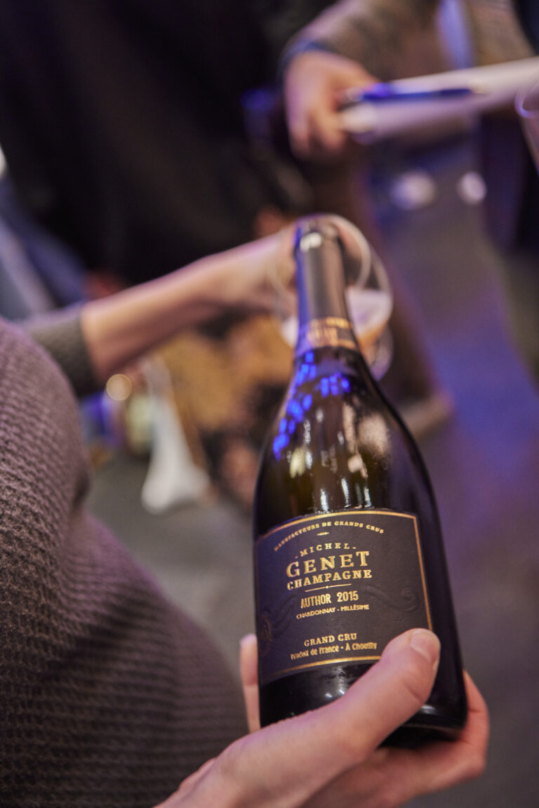 Eine Person hält eine Flasche Michel Genet Champagner bei einer Veranstaltung von Kirberg Catering Sachsenturm Köln, während sich die Gäste im unscharfen Hintergrund mischen.