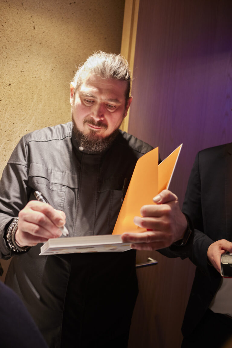 Ein bärtiger Mann mit Dutt unterschreibt Papiere, während er einen orangefarbenen Ordner im Kirberg Catering Sachsenturm Köln in der Hand hält.