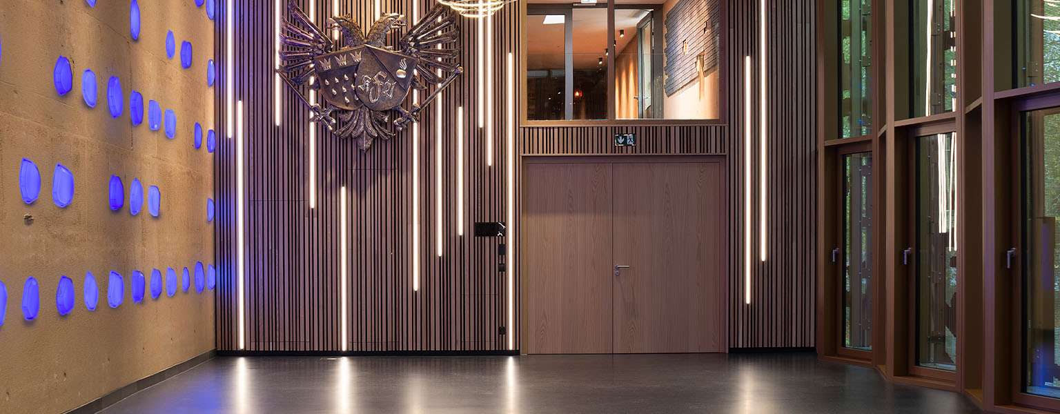 Eine moderne Lobby mit vertikalen Lichtbändern, blauen Wandleuchten, großen Fenstern und einem dekorativen Wandwappen empfängt die Gäste bei Kirberg Catering Sachsenturm Köln.