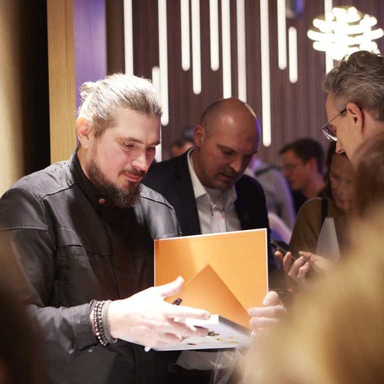 Ein Mann mit Bart und Pferdeschwanz hält eine orangefarbene Mappe in der Hand, während er in einer belebten Halle mit Menschen über Nachhaltigkeit im Catering diskutiert.